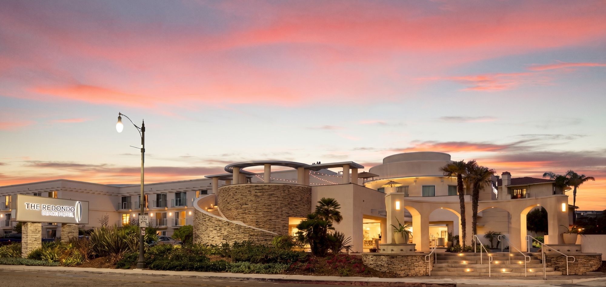 A modern, airy building with curved white walls sits by a palm-lined street at sunset, lit lamps glowing as pink and orange clouds fill the sky.