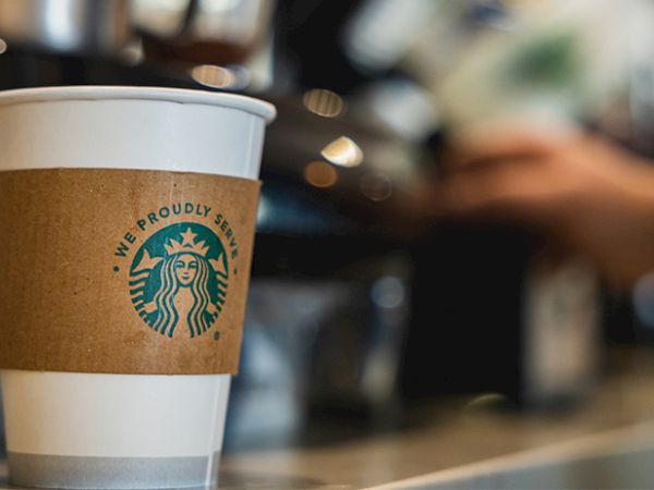 A white paper Starbucks cup with a brown sleeve sits at a counter, blurred barista in the background, cozy cafe vibe.
