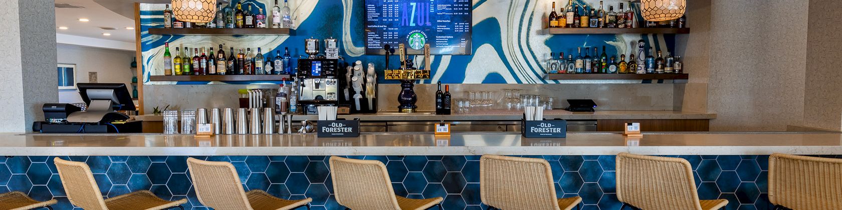 A stylish beach bar with a blue tiled front, eight wicker stools, a long counter, and bottles on glass shelves behind, under pendant lights. The scene is bright and modern.