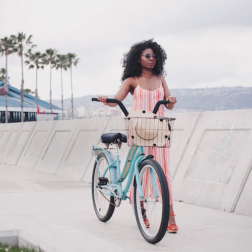 A woman with curly hair in a pink dress stands with a pastel bicycle by a sunny boardwalk, palm trees lining the path near the ocean.