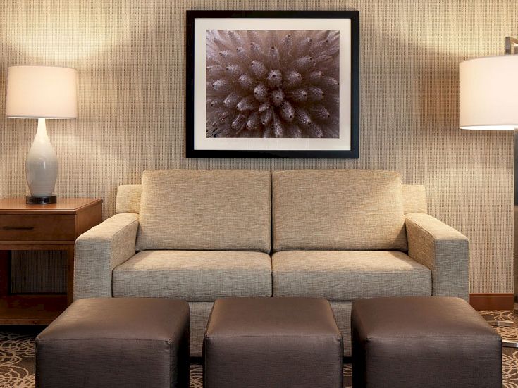 A simple living room with a beige sofa, two matching lamps, framed abstract art, and three brown ottomans in front of it.
