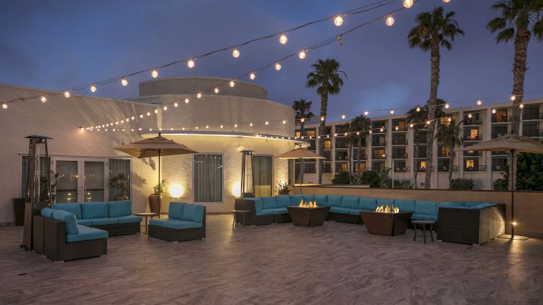A chic rooftop lounge with blue sectional seating, string lights, palm trees, and a curved white cabana at dusk, inviting relaxation.