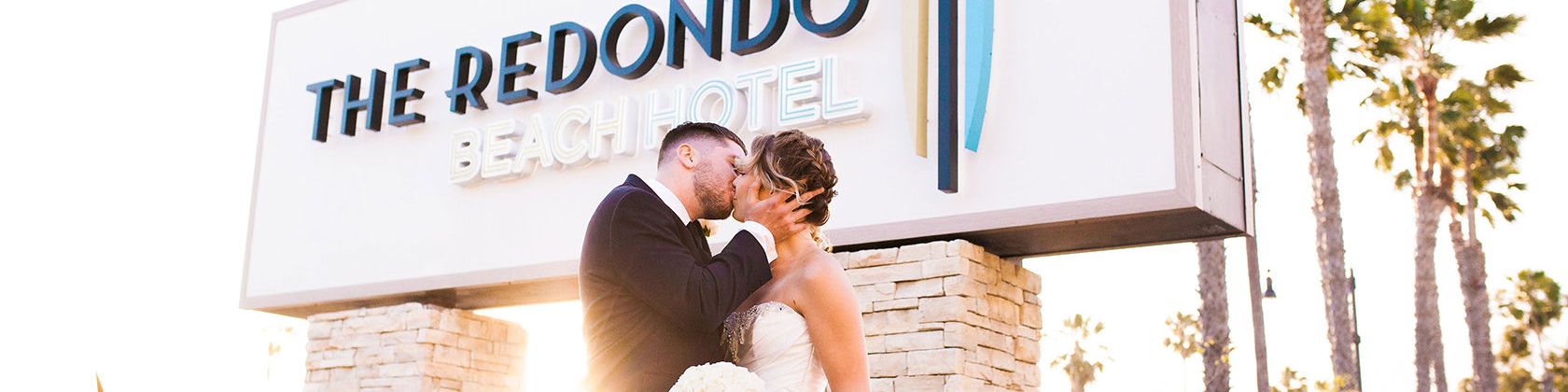 Two newlyweds kiss in front of The Redondo Beach Hotel sign, bride in a white gown holding a bouquet, sunny palm-lined backdrop.