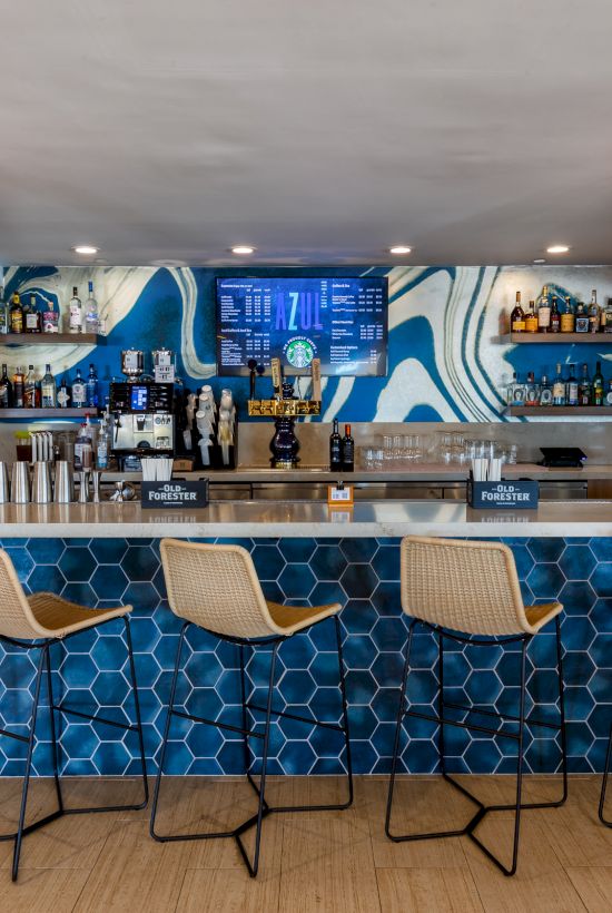 A modern bar with a blue hexagon-tiled front, high stools, bottled drinks, and hanging pendant lights behind the counter.