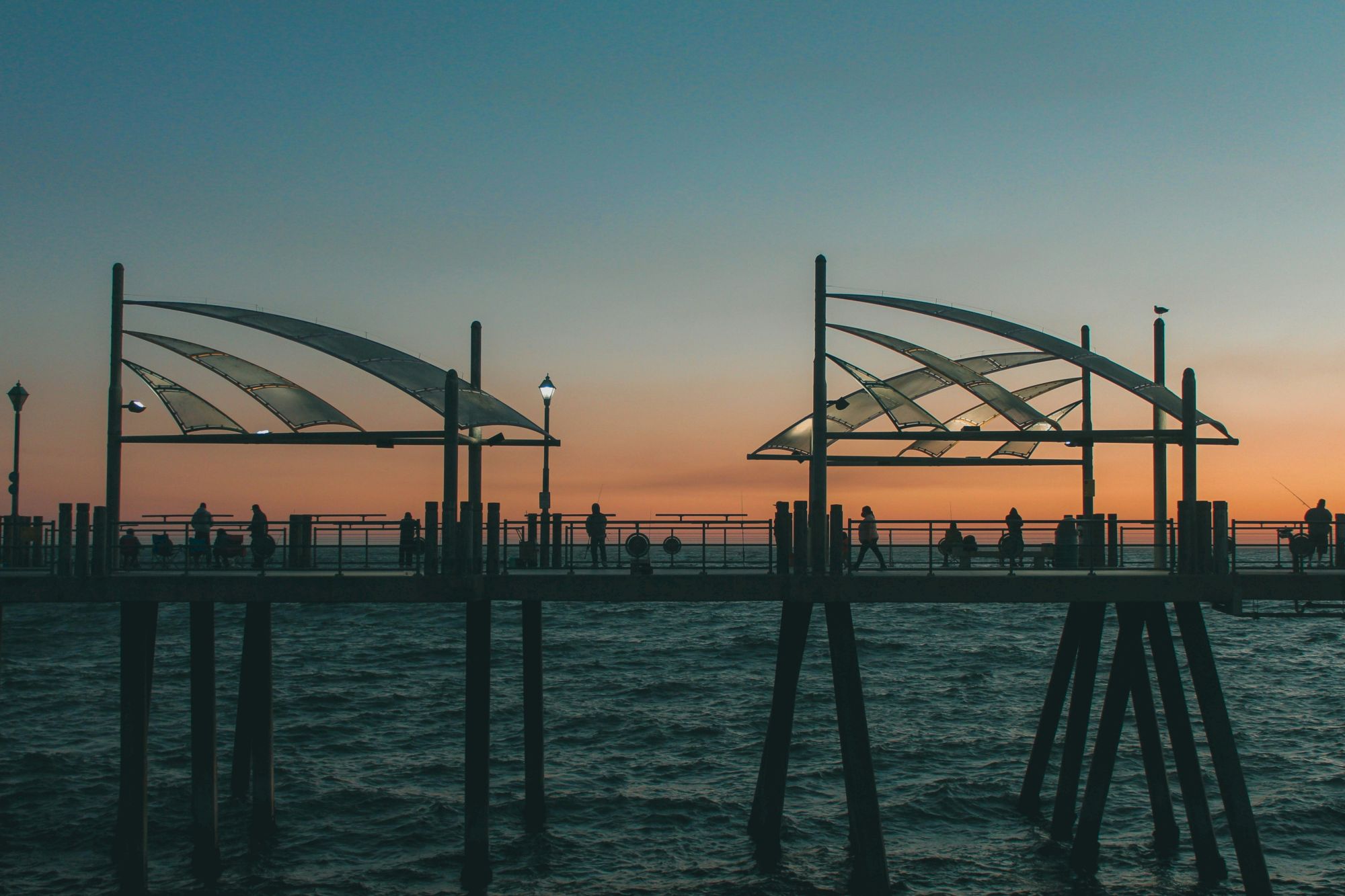 A wooden pier extends into calm water at sunset, with silhouetted people strolling under curved metallic canopies against a warm, orange-to-blue sky.