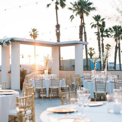 A chic outdoor wedding setup on a terrace with white tables, elegant chairs, glassware, and soft sunset light among palm trees.