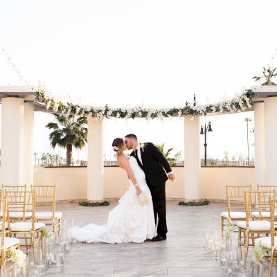 A bride and groom kiss under a floral arch at an outdoor wedding aisle, surrounded by gold chairs, bright daylight, and a celebratory mood.