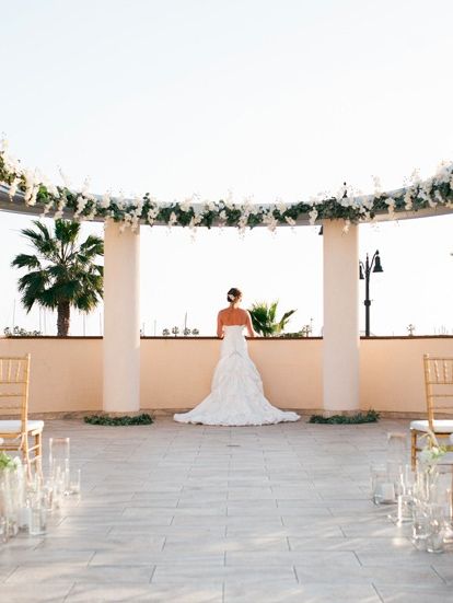 A bride in a white gown stands under a decorated arch at an outdoor wedding venue with rows of golden chairs and string lights.